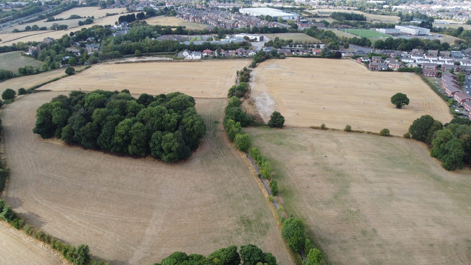Aerial view looking north across two harvested fields on the edge of Kingstone, with a wooded copse to one side, a track running between the fields, and the houses, schools and playing fields of Kingstone beyond, with Barnsley on the skyline.
